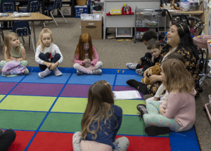 Kindergarten students listening to their teacher while sitting on a carpet